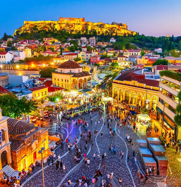 High angle view of a town in the evening, with lit up buildings. A rocky hill with ruins on the top.