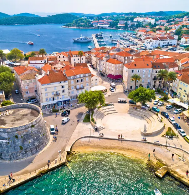 Aerial view of a town, with cream buildings with orange roofs, going into the distance on the right with, overlooking the sea on the left. In the forefront a circular stone building, and a circular seating area, surrounded by cars and some pedestrians. Slight view of water also in the forefront.