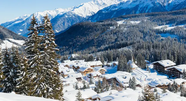 View of a village in the mountains in thick snow. With large fir trees in the left forefront, and smaller ones dotted around the village at the bottom the mountain. Above this a large forest of trees can be seen, going up the mountain, in front of large mountains covered in snow, in front of a light blue sky.