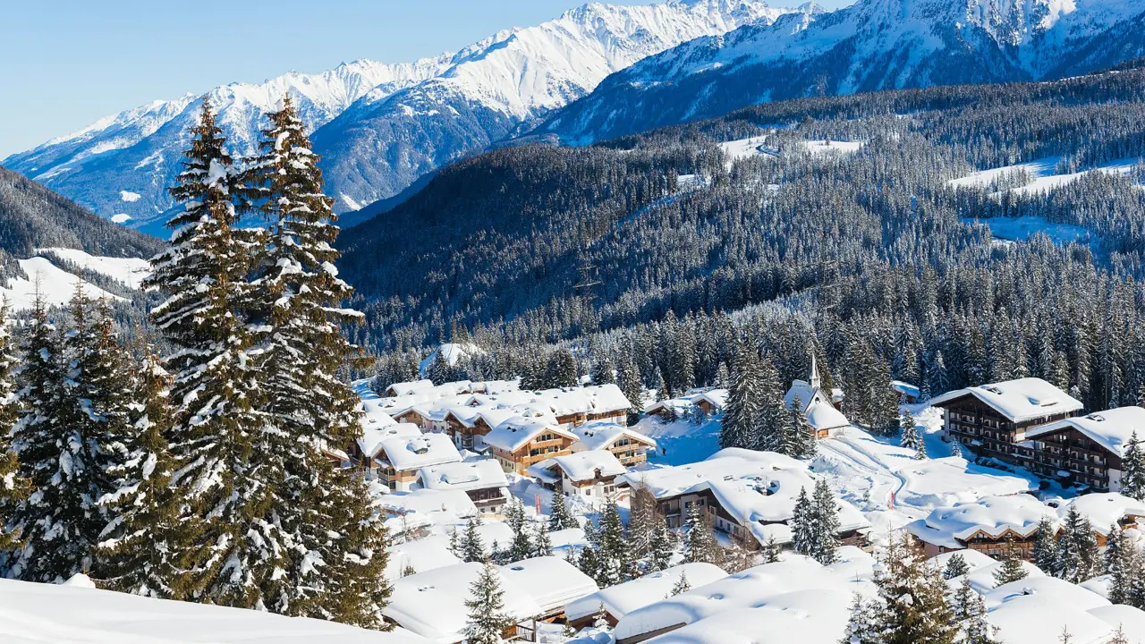 View of a village in the mountains in thick snow. With large fir trees in the left forefront, and smaller ones dotted around the village at the bottom the mountain. Above this a large forest of trees can be seen, going up the mountain, in front of large mountains covered in snow, in front of a light blue sky.