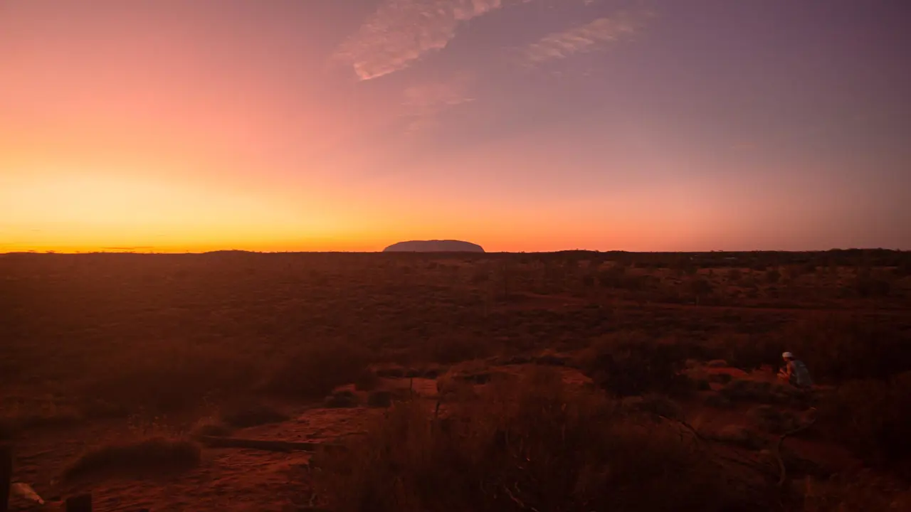 A massive red sandstone monolith rising out of the desert at Uluru in Australia, surrounded by sparse vegetation and open landscape.