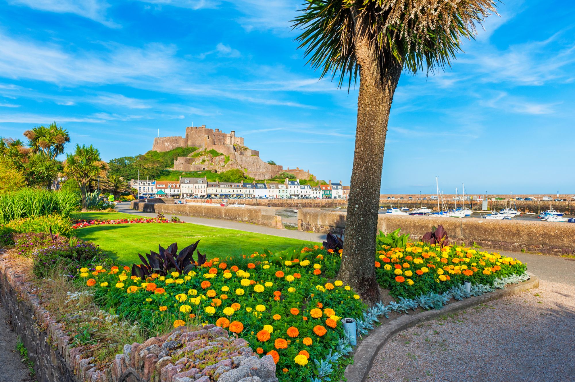 Long shot of Gorey Castle from the land, including a bed of orange and yellow flowers, a palm tree and grass in the forefront and the harbour and the houses next to it