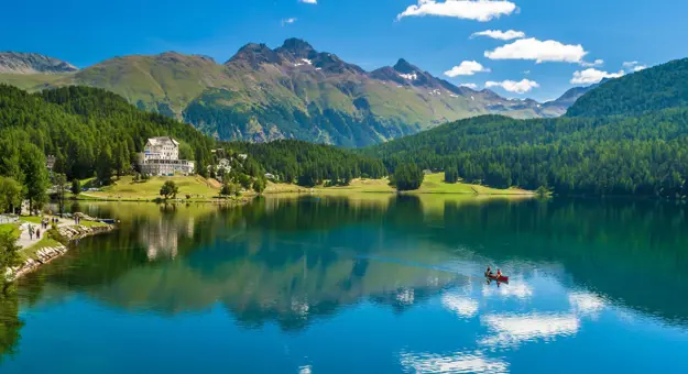 St Moritz Lake, Switzerland, showing a kayak travelling across the water, a large house on the bay and mountains in the background