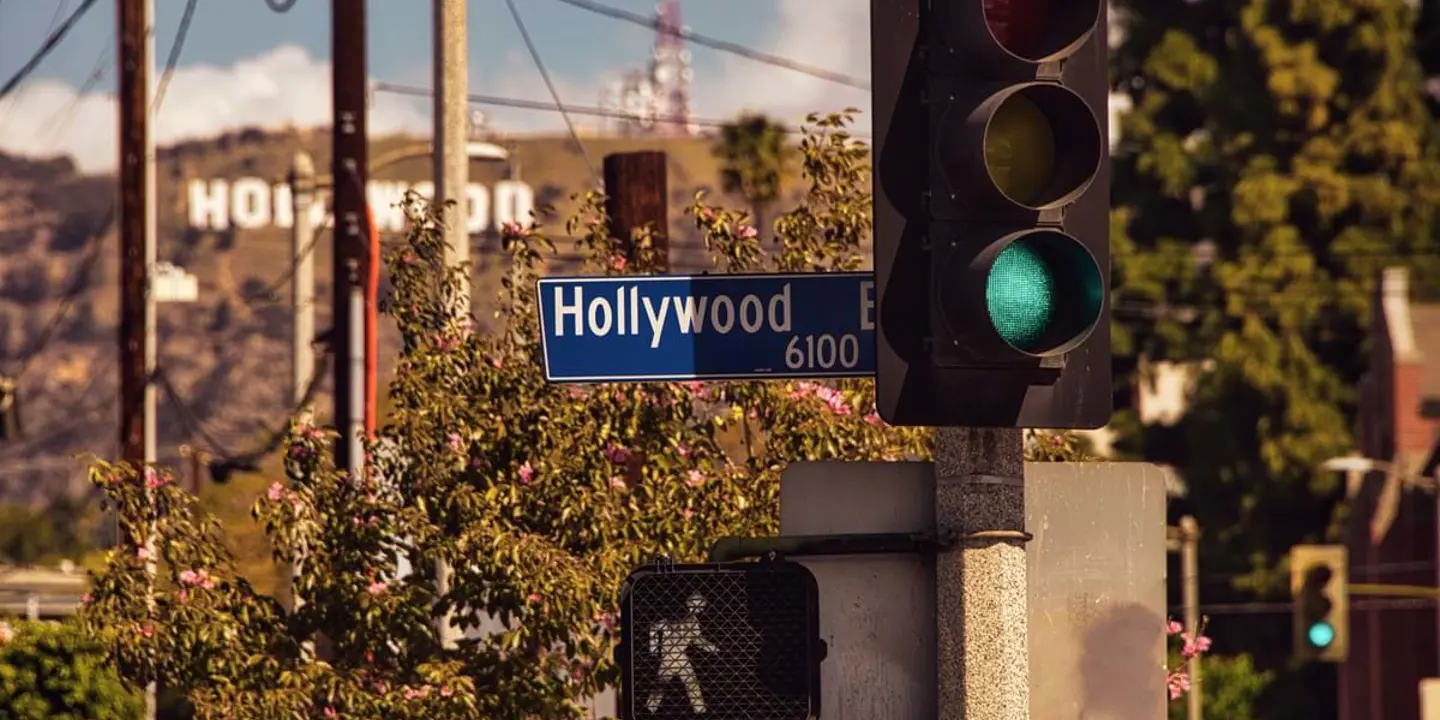 A Hollywood Boulevard road sign next to a traffic light and a white pedestrian crossing signal showing a walking man, with the Hollywood Sign visible in the background