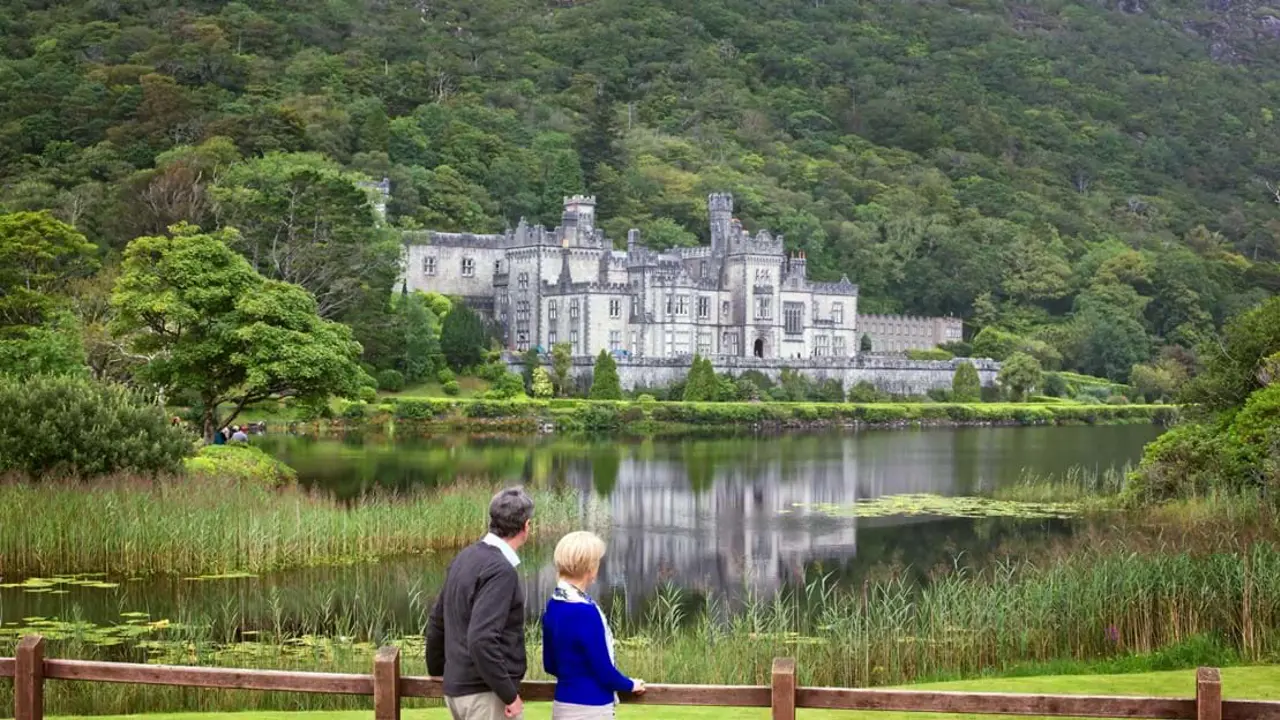 View of a grey abbey on the waterfront of a lake. Surrounded by trees. Back of two people in the forefront looking out at it