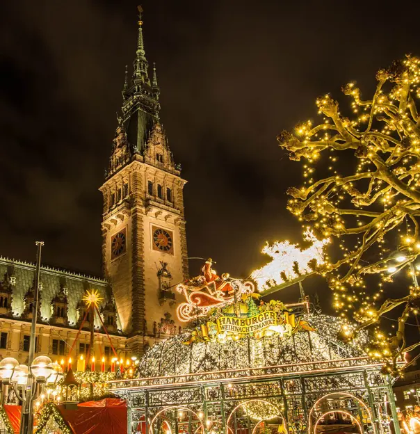 Low angle shot of a tall clock tower with spiked turret. Below, a sign for the Hamburg Christmas Markets against lots of fairy lights, with a lit up Santa Claus' sleigh and reindeers decoration, a tree to the right decorated with fairy lights, and artificial candles decoration to the left.