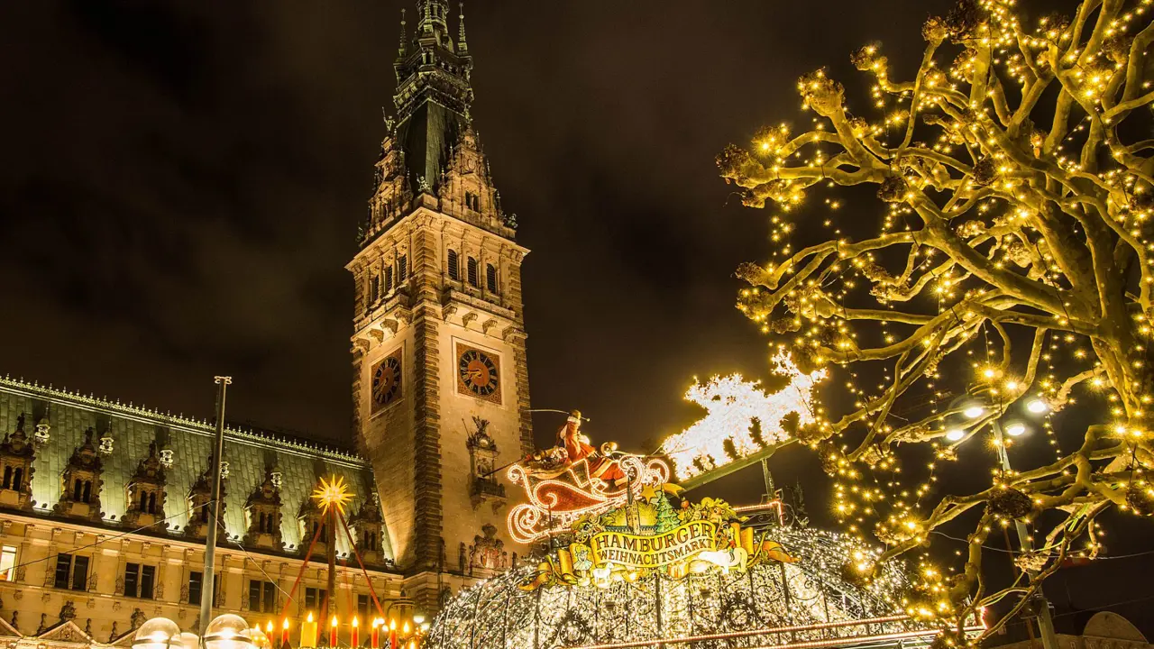 Low angle shot of a tall clock tower with spiked turret. Below, a sign for the Hamburg Christmas Markets against lots of fairy lights, with a lit up Santa Claus' sleigh and reindeers decoration, a tree to the right decorated with fairy lights, and artificial candles decoration to the left.