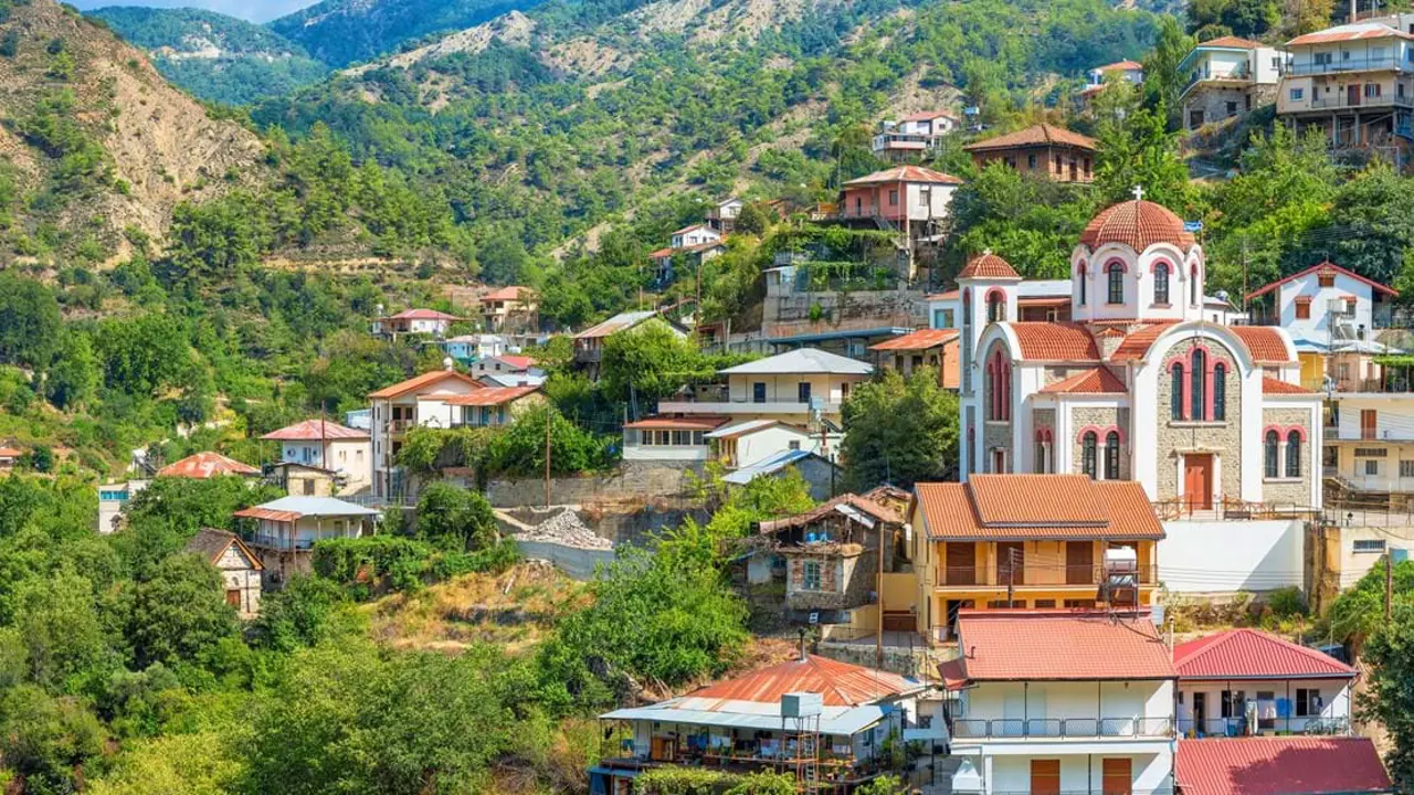 View of a town in the hills amongst the trees, with houses with orange and red roofs and a orange and white church to the right with curved roofs and a dome on top with a cross on. To the left is the hills behind, covered in trees.