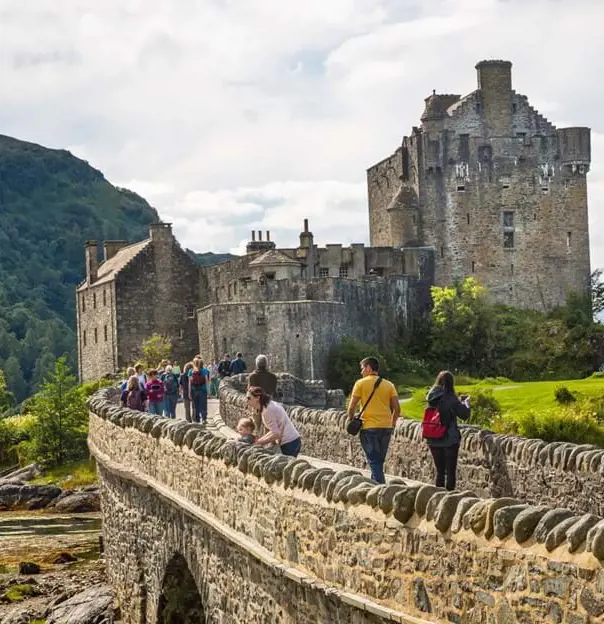 Eilean Donan Castle with people walking on the stone bridge walking towards it, and a mountain covered in trees behind it