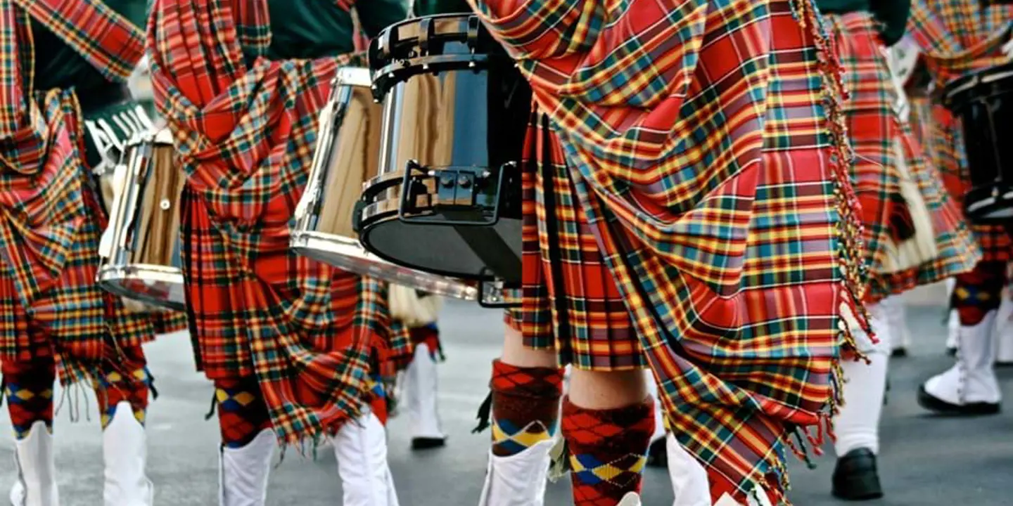 Close up of the bottom half of Scottish drum players in a parade wearing kilts, tartan shawls, kilt socks and white boots,holding drums 