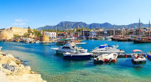 Boats docked at a harbour with bright blue water, buildings on the other side, and ruins of a castle to the left. Mountains in the distance in front of a blue sky.