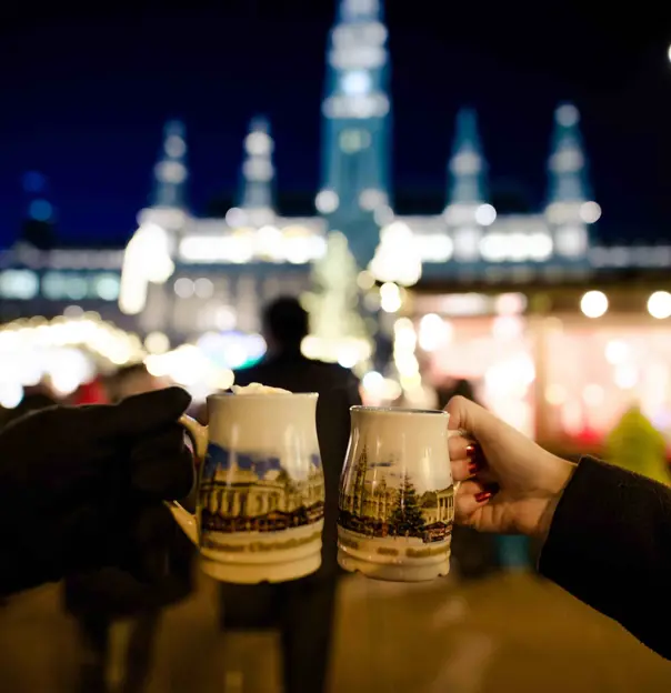 Two arms clinking two mugs of hot chocolate together in front of a blurry, lit up background