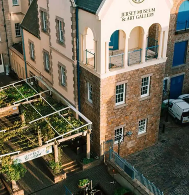 Bird's eye view of the Jersey Museum and Art Gallery, showing an open roof with plants tied to it and the Jersey Museum sign at the front of it to the left, and a car park with some parked white vehicles to the right