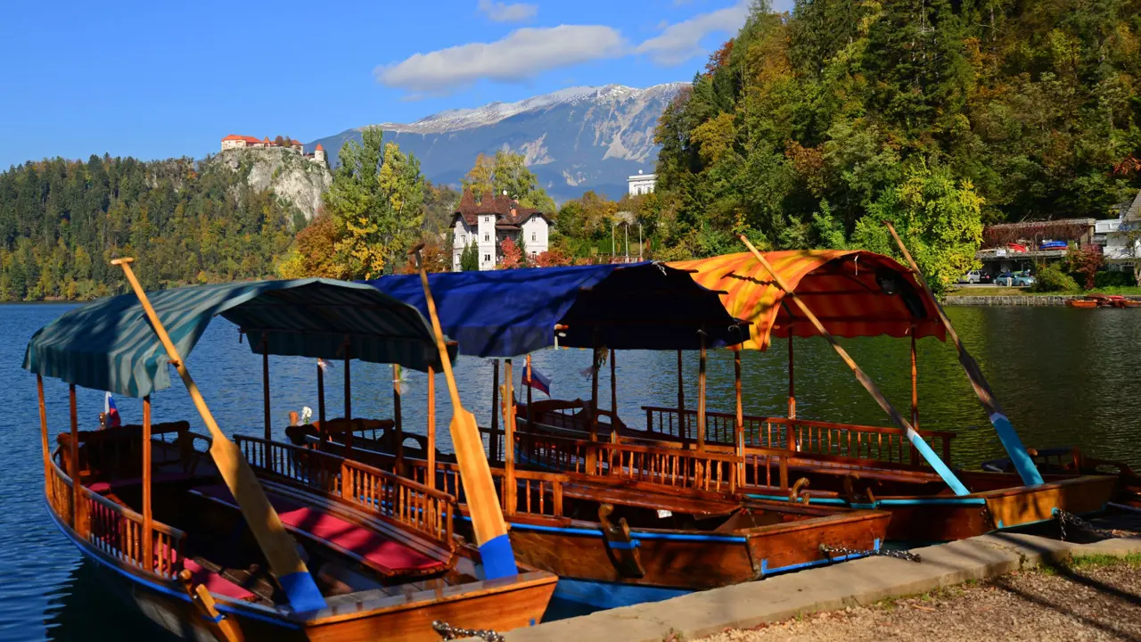 Back of two pletna boats docked on Lake Bled, with forests on the other side. A large building on a hill and one on ground level, and mountains in the distance.