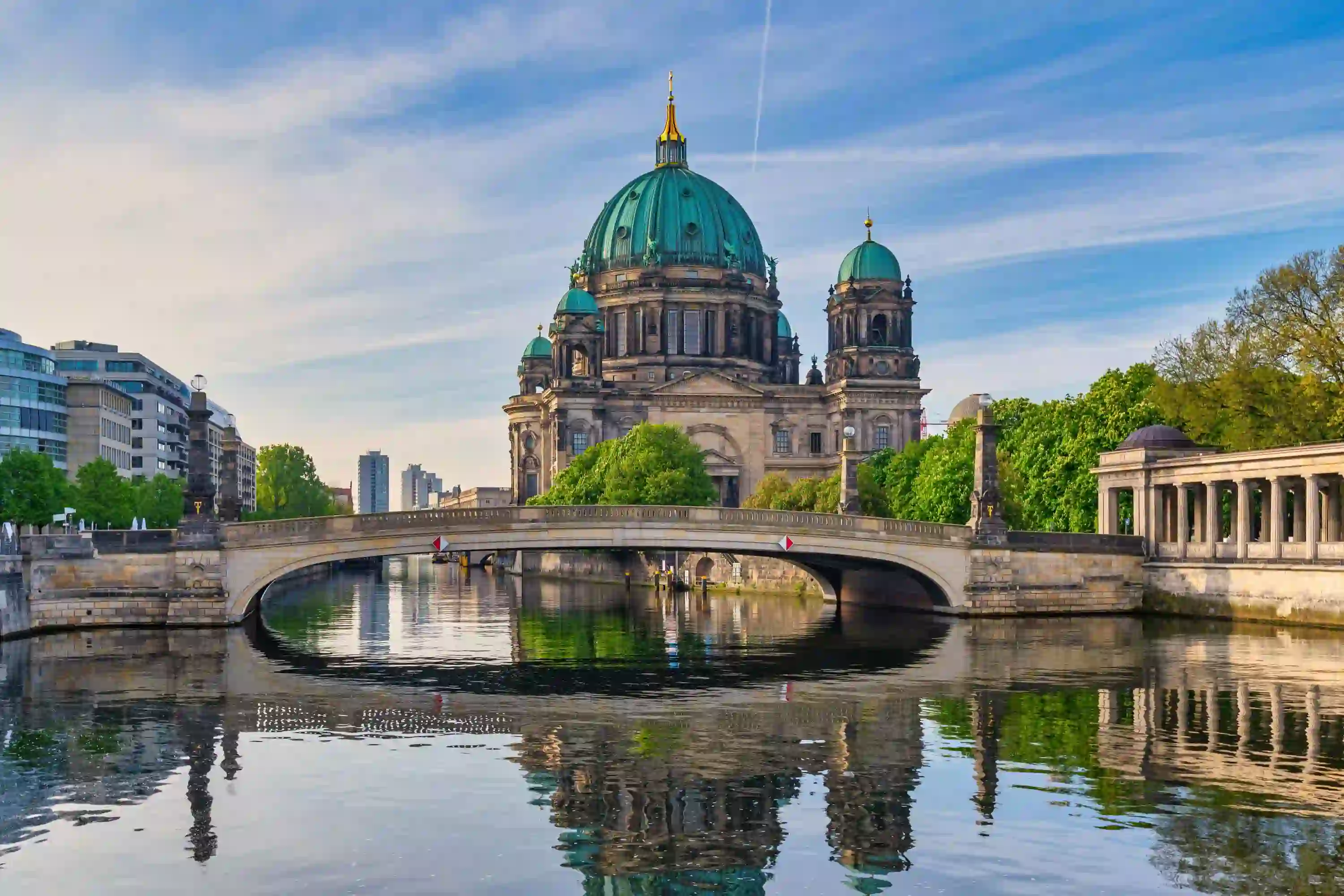 View of Berlin Cathedral, which is grey with turquoise domed roofs, and has a gold turret in the middle. In front is a bridge, and there is water in the forefront