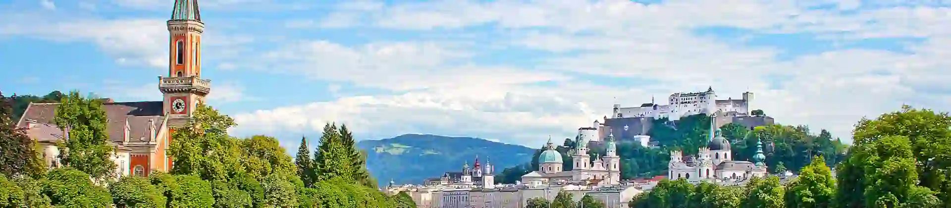 View of a river winding to the left, with grass and trees on either side, and a red church with a tall, slim tower with a blue turret. On the left of the river multiple large buildings can be seen, with numerous turquoise turrets pointing out, and the fortress on the top of the hill, all under a blue sky