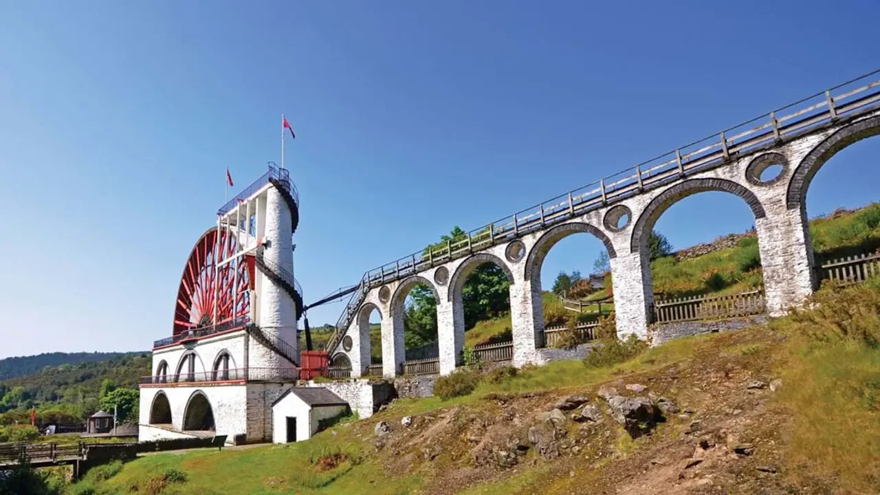 Low angle shot of the Laxey Wheel in the Isle of Man, which has a white base with a red wheel, on a grassy hill in front of a blue sky 