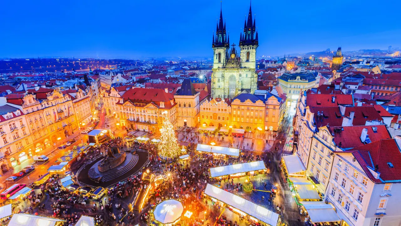 High angle shot of Christmas market in the town square of Prague, showing a lit up gothic church with two towers and spiky turrets, a monument with a circular barrier around it in the centre of the square, next to a large lit up Christmas tree. White roofs of the stalls are all around the square surrounded by people. 