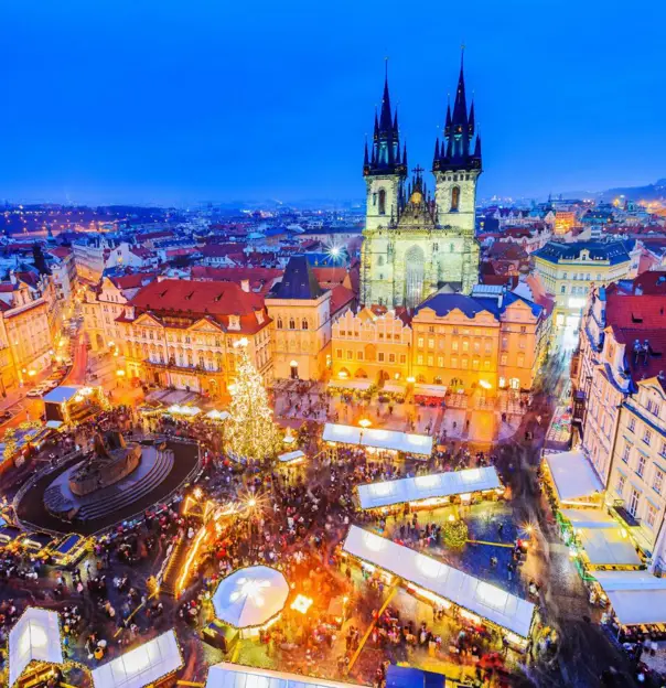 High angle shot of a Christmas market in the town square of Prague, showing a lit up gothic church with two towers and a large lit up Christmas tree