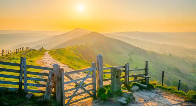 View of the Peak District at sunrise, with a gate at the forefront of the image and a trail that leads all the way to the mountains in the far distance