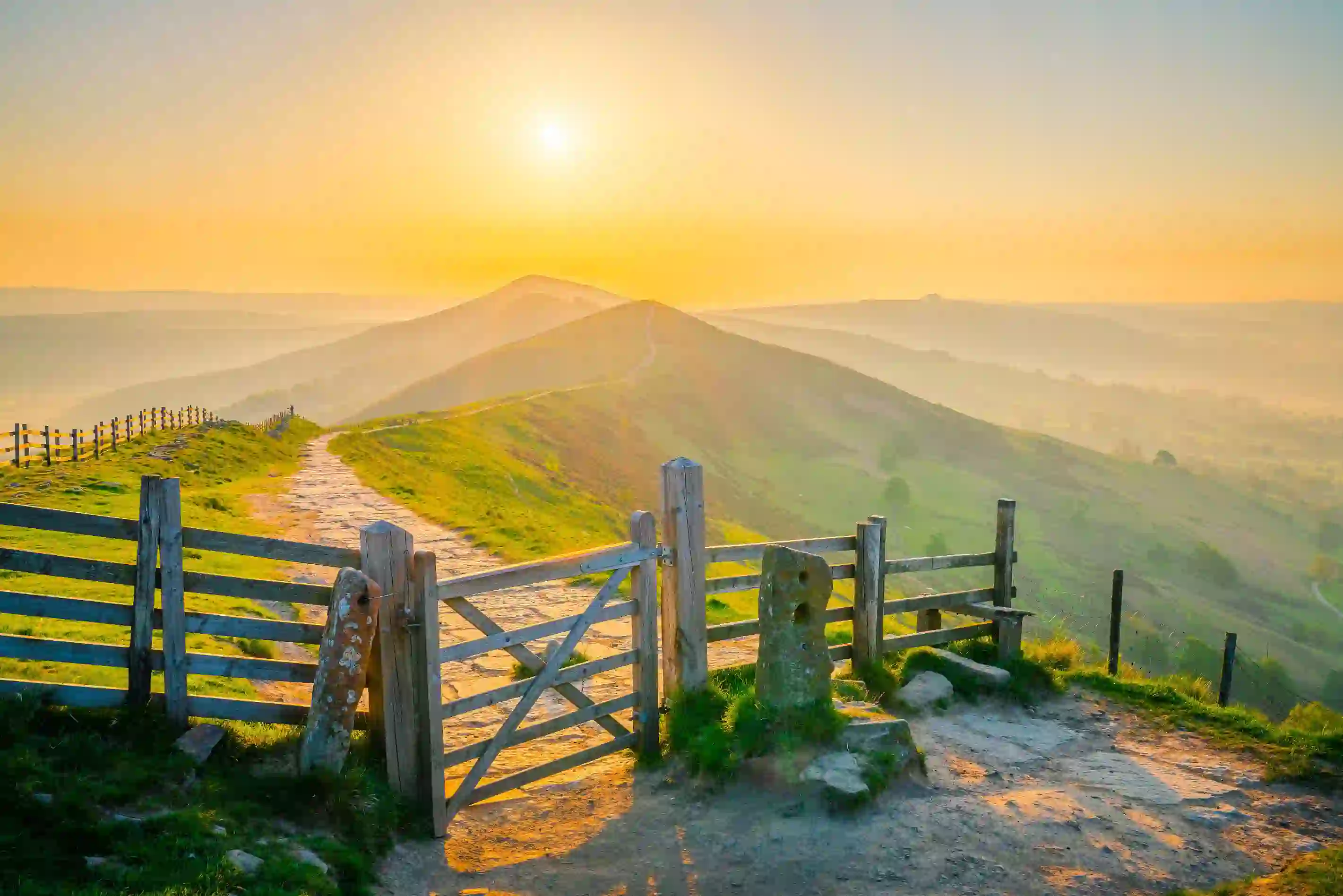 View of the Peak District at sunrise, with a gate at the forefront of the image and a trail that leads all the way to the mountains in the far distance