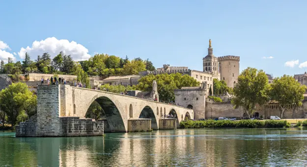 Wide angle shot of the Pont d'Avignon bridge, with the palace on the other end. The river water in the forefront, and a blue sky with clouds above.