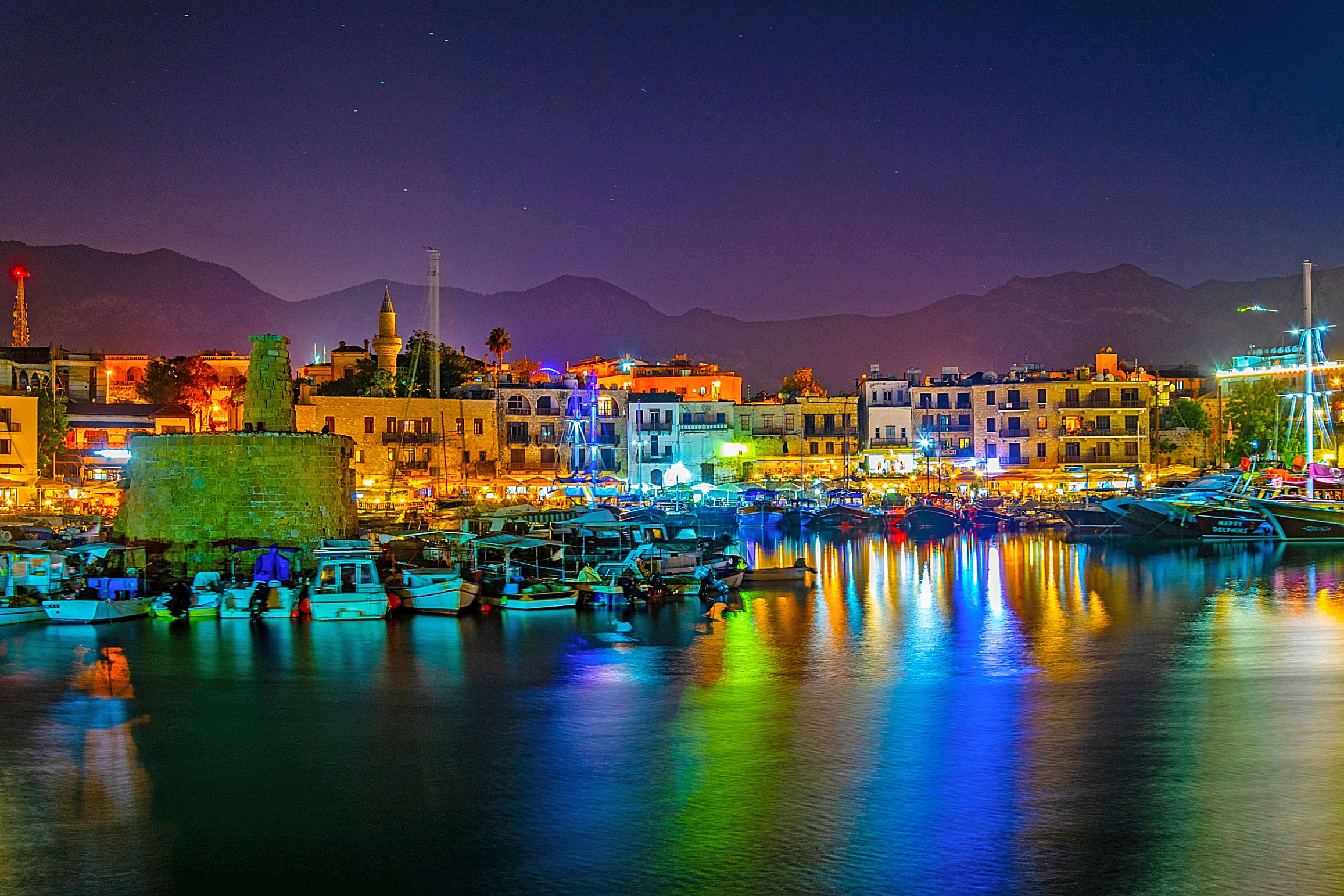 View of a harbour at night, with boats docked and a ruin to the left. Multi coloured lights behind the boats, reflecting on the water. A strip of buildings on the far side of the harbour, in front of a silhouette of mountains in front of a night sky.