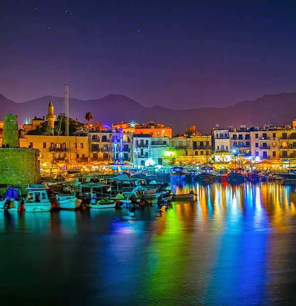 View of a harbour at night, with boats docked and a ruin to the left. Multi coloured lights behind the boats, reflecting on the water. A strip of buildings on the far side of the harbour, in front of a silhouette of mountains in front of a night sky.