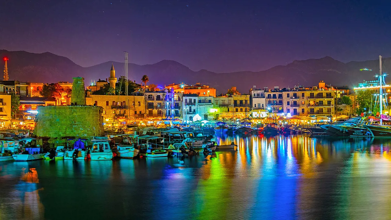 View of a harbour at night, with boats docked and a ruin to the left. Multi coloured lights behind the boats, reflecting on the water. A strip of buildings on the far side of the harbour, in front of a silhouette of mountains in front of a night sky.