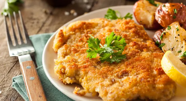 Close up of half a plate of food, with a shnitzel and a lemon and some potatoes next to it. A fork to the left of the dish with a turquoise napkin underneath it.