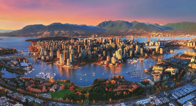 Aerial view of Vancouver, Canada at sunset, showing the city skyline, surrounding water, and distant mountains beneath a glowing, colourful sky