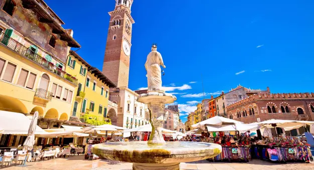 View of Fontana Madonna fountain from behind, showing the Torre dei Lamberti tower and the square's restaurants and markets