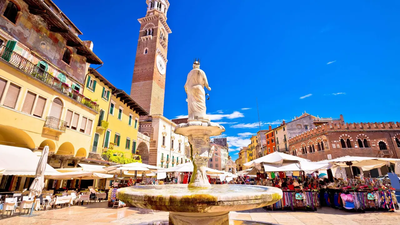 View of Fontana Madonna fountain from behind, showing the Torre dei Lamberti tower and the square's restaurants and markets