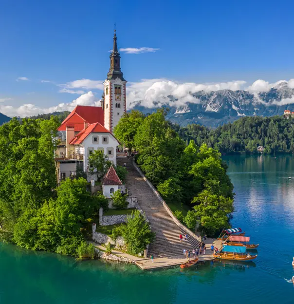 Shot of a church on a small bit of forested land on water, there is a pathway leading up to the church which has some people and boats at the bottom. Behind, there are mountains and clouds, under a blue sky. 
