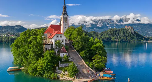 Shot of a church on a small bit of forested land on water, there is a pathway leading up to the church which has some people and boats at the bottom. Behind, there are mountains and clouds, under a blue sky.
