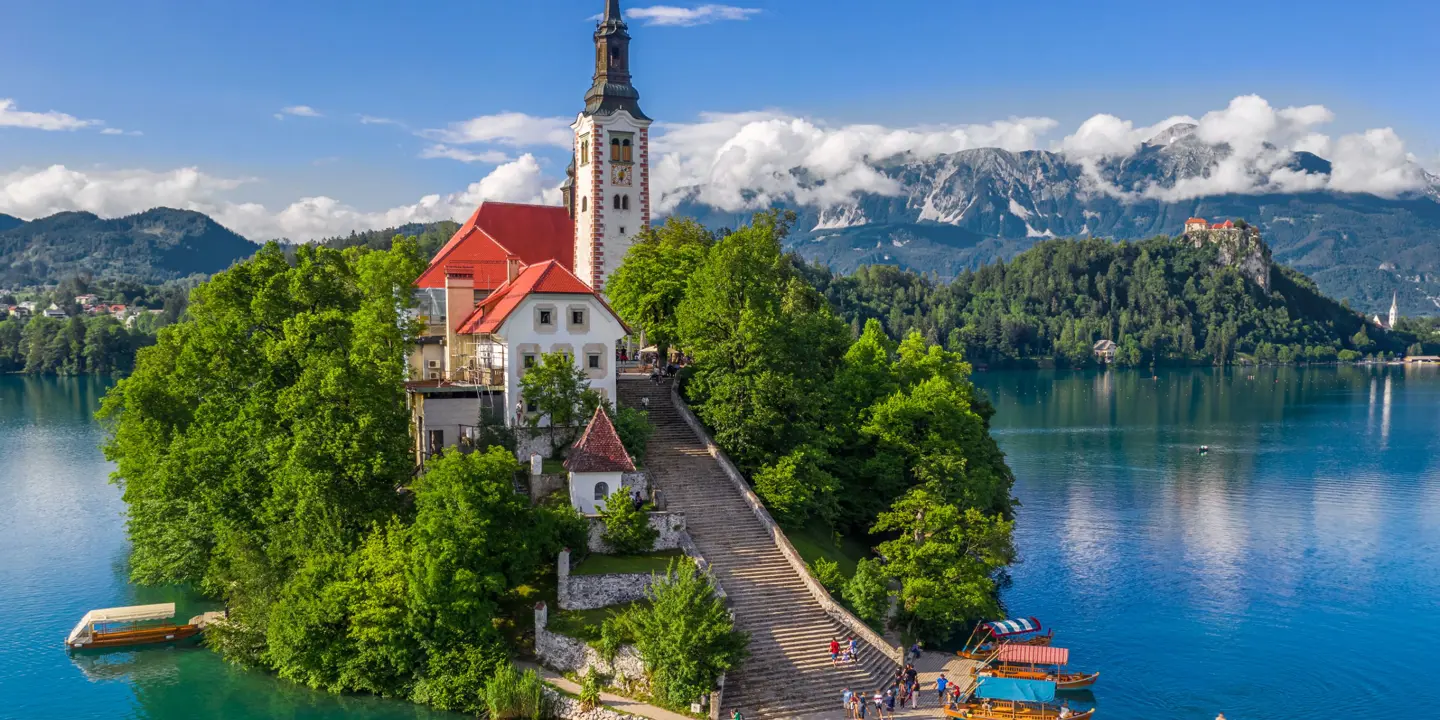 Shot of a church on a small bit of forested land on water, there is a pathway leading up to the church which has some people and boats at the bottom. Behind, there are mountains and clouds, under a blue sky.