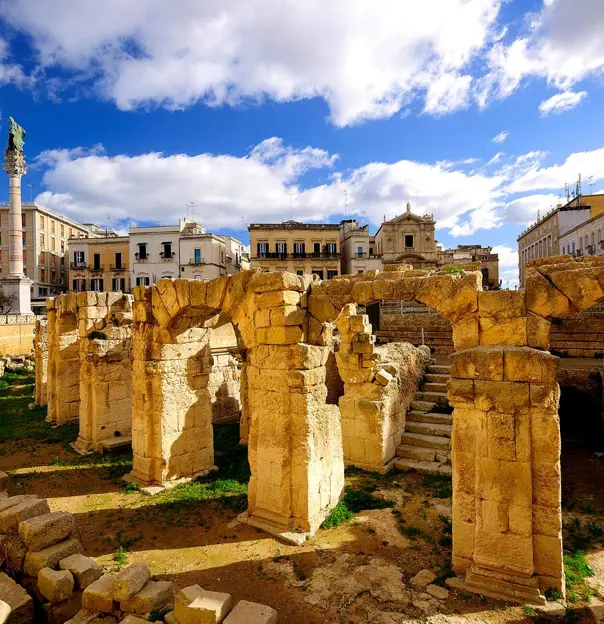 Ancient Roman ruins in the city centre of Lecce, Puglia, Italy, with weathered stone arches and columns surrounded by historic buildings under a bright blue sky with scattered clouds