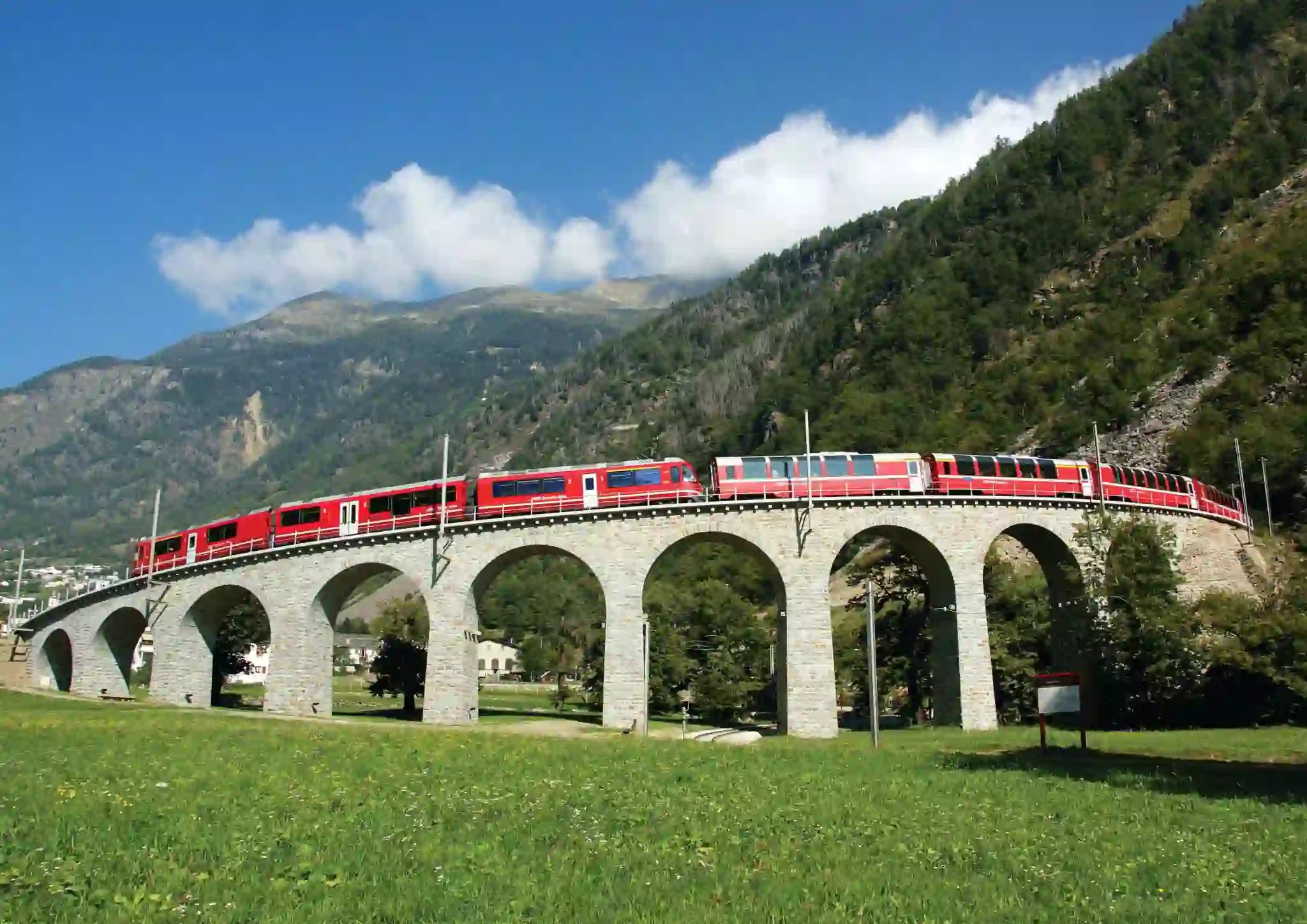 The Bernina Express going across a bridge, with grassy mountains in the background