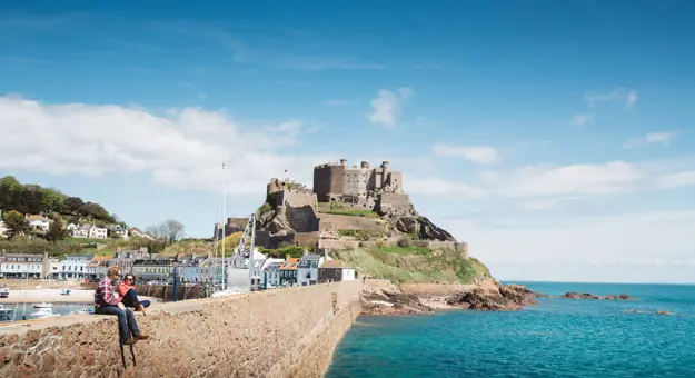 Long shot of Mont Orgueil Castle, showing part of the harbour below it and two people sat on the wall between the harbour and the sea, the buildings beside the harbour and the houses on a hill behind it, on a sunny day with a blue sky 