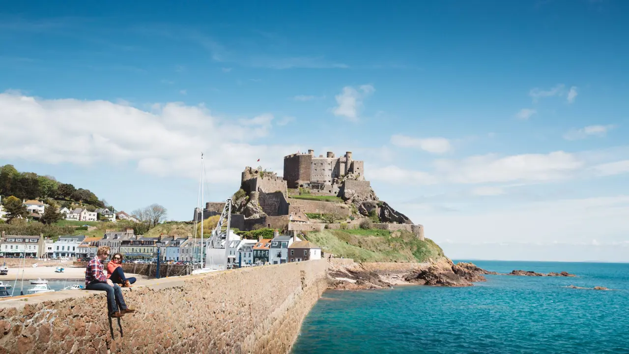 Long shot of Mont Orgueil Castle, showing part of the harbour below it and two people sat on the wall between the harbour and the sea, the buildings beside the harbour and the houses on a hill behind it, on a sunny day with a blue sky 