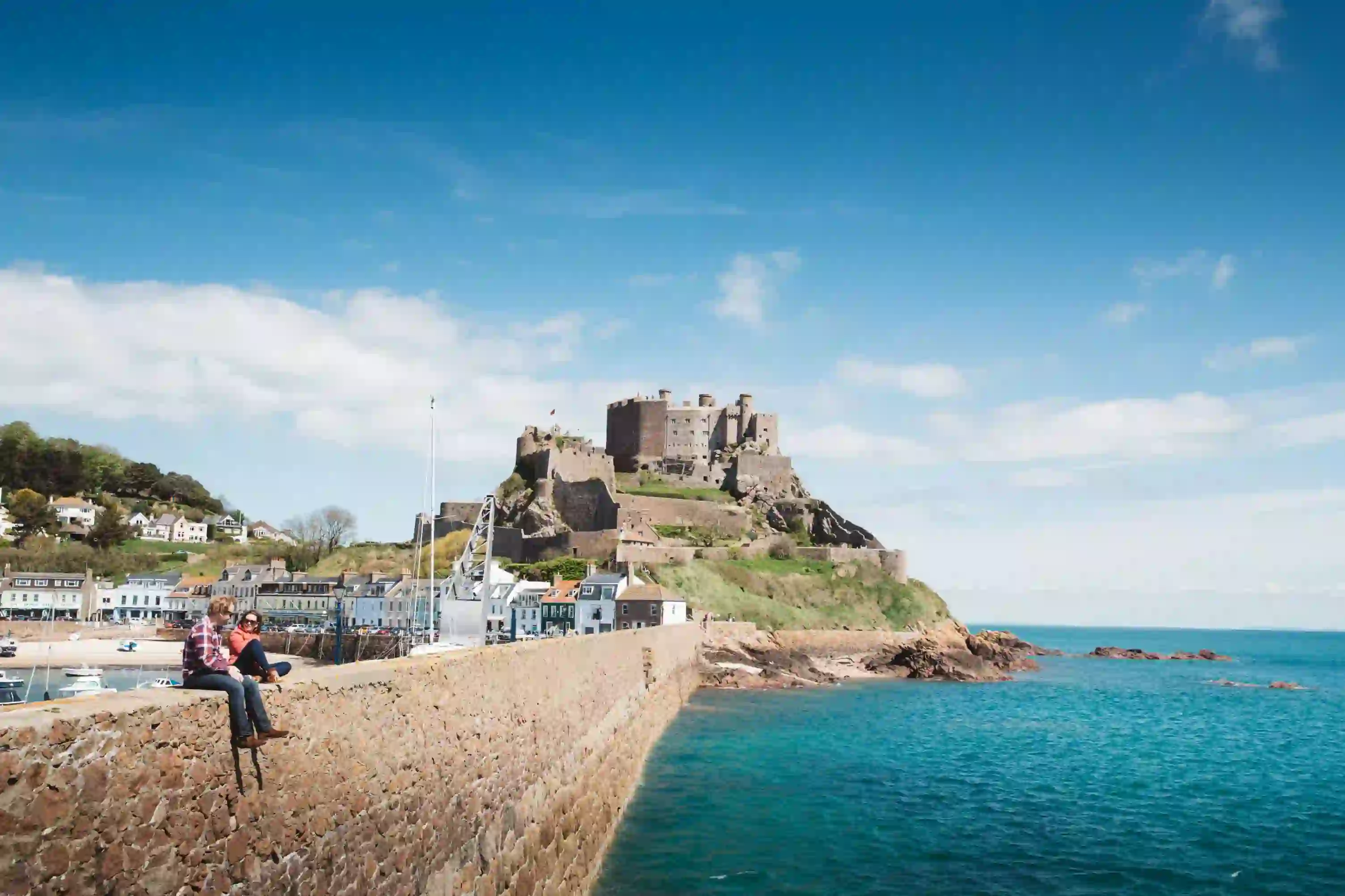 Long shot of Mont Orgueil Castle, showing part of the harbour below it and two people sat on the wall between the harbour and the sea, the buildings beside the harbour and the houses on a hill behind it, on a sunny day with a blue sky 