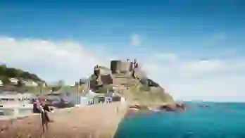 Long shot of Mont Orgueil Castle, showing part of the harbour below it and two people sat on the wall between the harbour and the sea, the buildings beside the harbour and the houses on a hill behind it, on a sunny day with a blue sky
