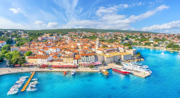 High angle shot of a town on the coast, with bright blue water and boats docked to the left. The town is full of buildings with orange roofs, and the distance is forested land. The sky is blue with some clouds. 