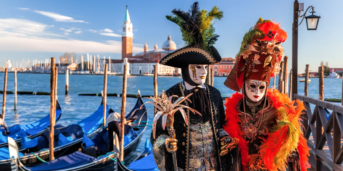 Two people with masks and fancy dress standing in front of boats, a body of water and the city of Venice