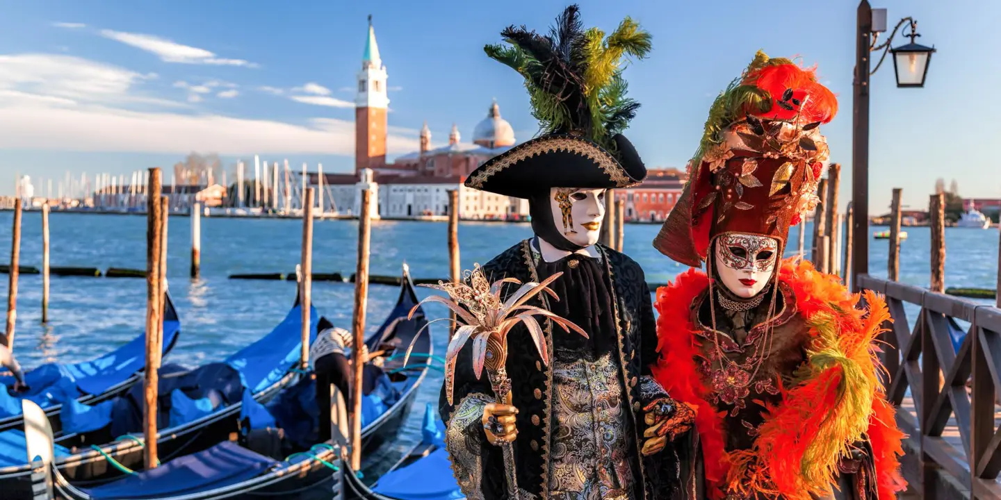 Two people in elaborate Venetian carnival costumes and masks standing by gondolas on a canal, with the Church of San Giorgio Maggiore in the background