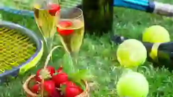 Close up of two glasses of champagne, a basket of strawberries, tennis balls and rackets on some grass