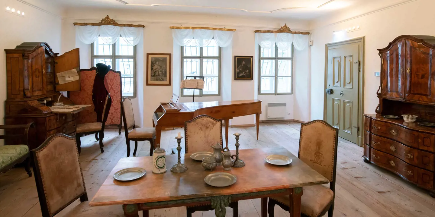 Inside a room with an antique wooden table in the front, with silver plates and two candle holders in the middle, three cream chairs surround the table. Behind is a wooden piano, and another chair at it. A sage door to the right, forwards, a dark wooden cabinet with drawers underneath. To the left, there is another one of these, with the doors open and a desk attached with paperwork on and another chair in front. The back wall has three grid windows and mesh white curtains and paintings between the windows.