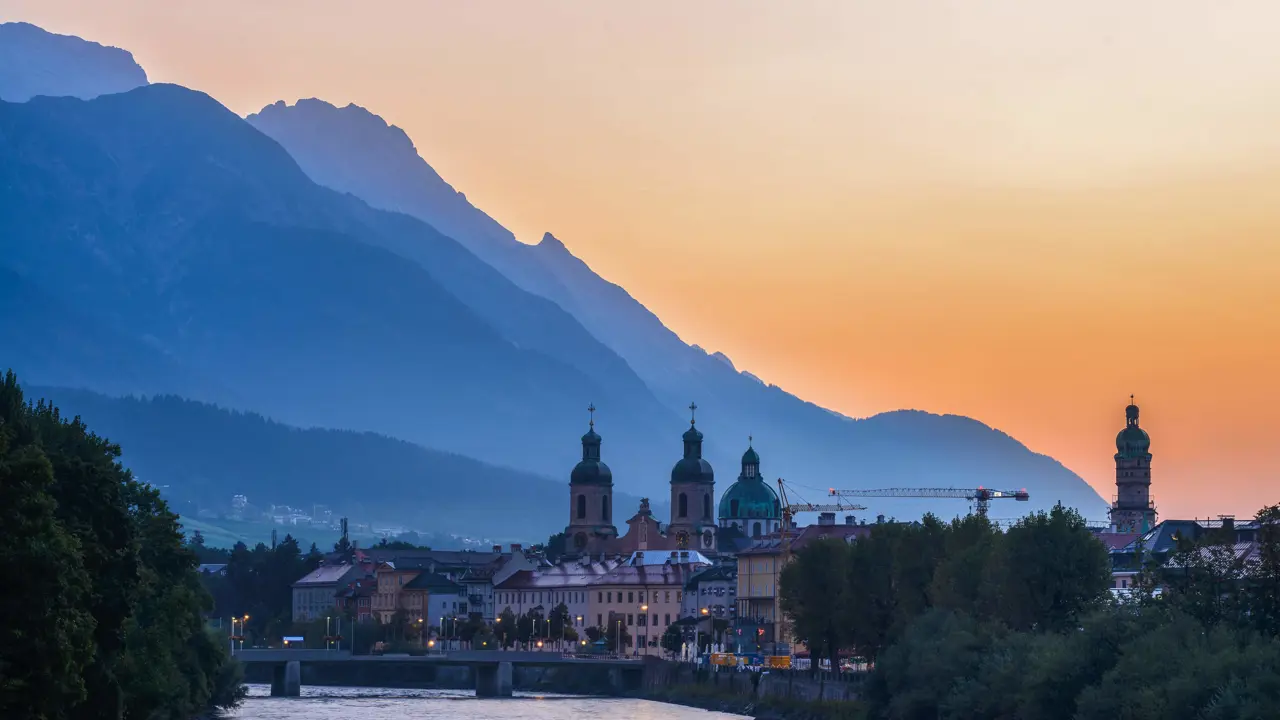 View of the town of Innsbruck at sunset from the river, with a bridge in the forefront and the town's buildings along the waterfront. Two clock towers, one dome shaped building and one tower poke out of the top of the cityscape. The silhouette of a mountain is behind, against an orange sky.