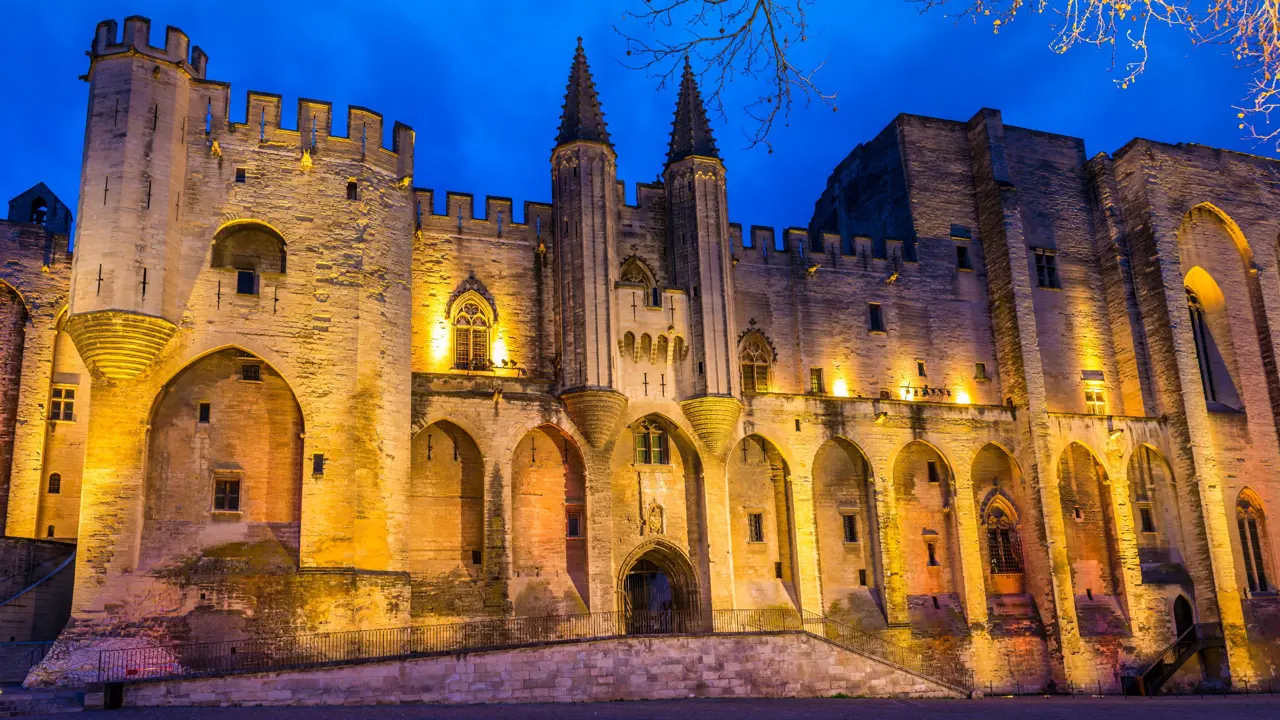 Low angle shot of a beige coloured stone palace, lit up with amber lights on the front. With pointy turrets in the middle and battlements on the left side. In front of a bright blue night sky.