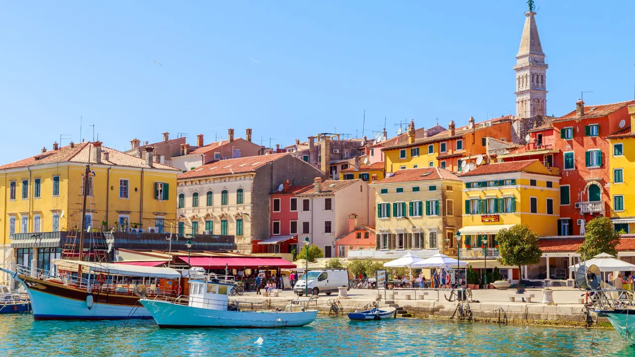 View of Rovinj from the water, showing boats docked at the harbour and colourful buildings. Slightly further back is a stone tower. The sky is blue and clear.